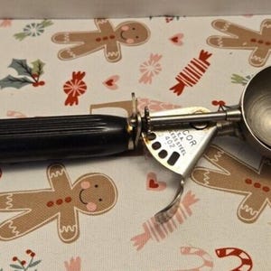 May include: A stainless steel ice cream scoop with a black fluted handle and a spring-loaded release mechanism. The metal frame is stamped with the text 'DECOR U.S.A. STAINLESS STEEL 402'. The scoop features a circular bowl and a side-mounted lever.
