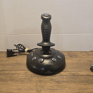 May include: A black Raider Pro joystick with a central vertical handle, a top button, and a circular base featuring four grey buttons, a side slider, and a connector cable. The base displays the text Raider Pro and Performance in white lettering.