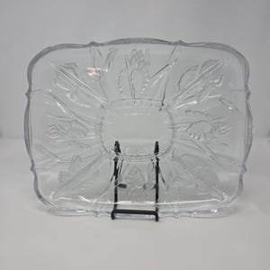 May include: A rectangular clear glass serving tray featuring a molded floral pattern with raised iris motifs. The tray has scalloped edges and a central oval section, providing a decorative surface for serving food items in a clear, transparent finish.