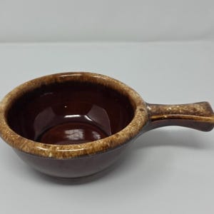 May include: A small, round, dark brown ceramic bowl with a single handle extending from the side. The bowl features a glossy finish with a lighter, mottled tan and cream-colored glaze along the thick, rounded rim and the top of the handle.
