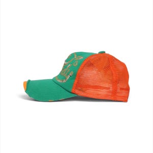 May include: A green and orange trucker cap featuring a mesh back panel and a solid front panel. The front displays a decorative gold-toned script design with small embellishments. The brim is green with a small orange accent on the edge.