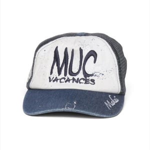May include: A trucker hat with a dark blue denim brim and a white front panel featuring the dark blue embroidered text MUC VACANCES. The hat has a dark blue mesh back and a small white embroidered signature on the right side of the brim.