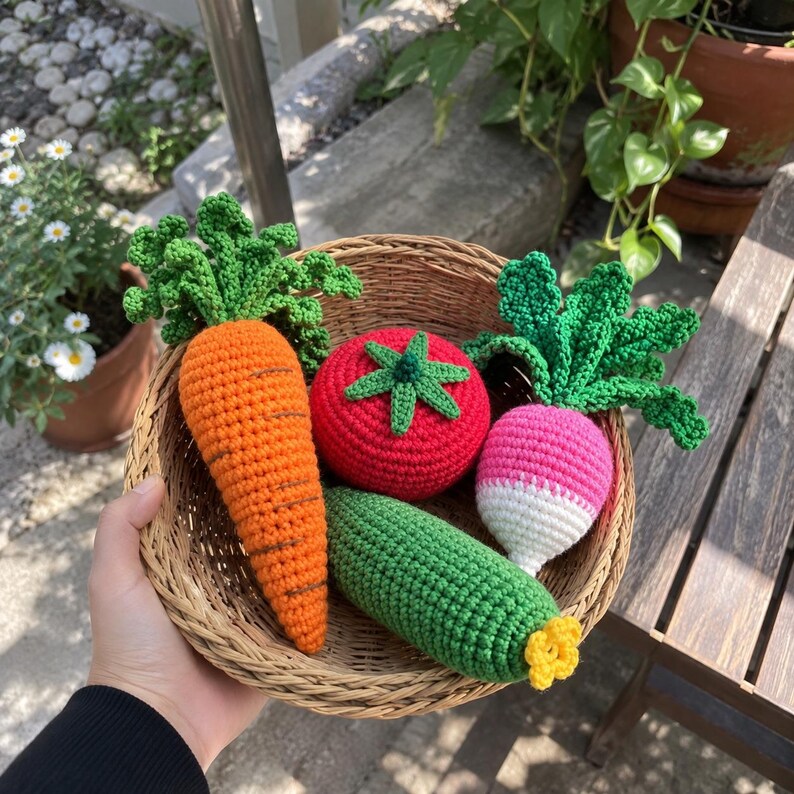 May include: A woven wicker basket containing four crocheted vegetables: a bright orange carrot with green leafy tops, a round red tomato with a green stem, a green cucumber with a yellow blossom end, and a pink and white radish with green leaves.