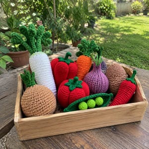 May include: A collection of crocheted toy vegetables in a wooden crate, including a white radish, red bell pepper, orange carrot, purple onion, brown potato, tan onion, red tomato, green pea pod with three peas, and a red chilli pepper.