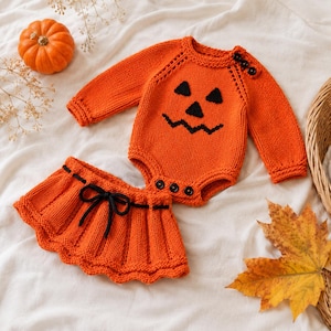 May include: An orange knit long-sleeve bodysuit featuring a black jack-o'-lantern face design with three black buttons at the shoulder and three at the crotch, paired with a matching orange knit skirt featuring a black drawstring tie at the waist.