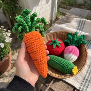 May include: A hand-crocheted orange carrot with green leafy tops held in front of a woven basket containing a red tomato, a green cucumber with a yellow tip, and a pink and white radish with green leaves, all crafted from yarn.