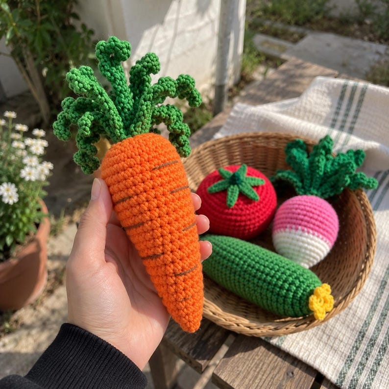 May include: A hand-crocheted orange carrot with green leafy tops held in front of a woven basket containing a red tomato, a green cucumber with a yellow tip, and a pink and white radish with green leaves, all crafted from yarn.
