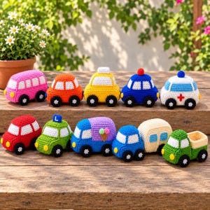 Puede incluir: Una colección de diez pequeños vehículos de juguete tejidos a ganchillo a mano, dispuestos sobre una superficie de madera. El conjunto incluye un autobús rosa, un coche naranja, un taxi amarillo, un coche de policía azul, una ambulancia blanca, un coche rojo, un taxi verde, un camión de helados morado, un coche azul y un camión verde.