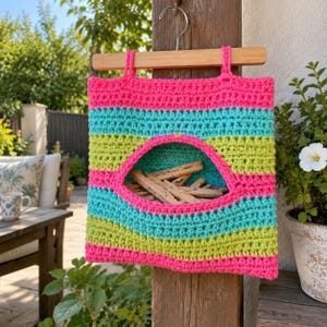 May include: A square crochet bag featuring horizontal stripes in neon pink, turquoise, and lime green. The bag has a large, central oval opening and is suspended from a wooden hanger. Inside the bag are several wooden clothespins.