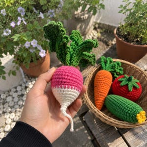 May include: A handcrafted crochet radish with a bright pink top, white bottom, and textured green leaves. Beside it, a woven basket contains a crocheted orange carrot, a red tomato with a green stem, and a green cucumber with a yellow flower.