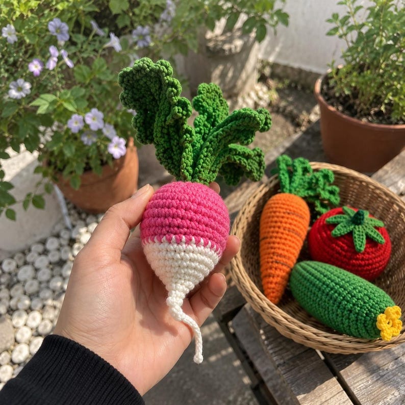 May include: A handcrafted crochet radish with a bright pink top, white bottom, and textured green leaves. Beside it, a woven basket contains a crocheted orange carrot, a red tomato with a green stem, and a green cucumber with a yellow flower.