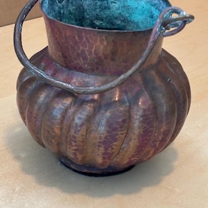 May include: A round, hammered copper bucket with a wide, fluted body and a dark, oxidized patina. The interior features a bright teal-green verdigris finish. A thick, curved copper handle is attached to the top rim with two metal loops.