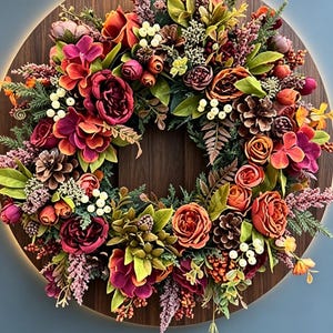 May include: A circular wreath featuring a mix of artificial flowers in shades of deep red, orange, and burgundy, interspersed with green foliage, small white berries, and brown pinecones, all mounted on a circular dark wood-grain base.