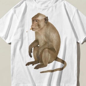 May include: A white short-sleeved t-shirt featuring a central graphic illustration of a brown monkey sitting in profile. The monkey is depicted with a small, light-coloured object held near its mouth, resembling a cigarette, with a tiny smoke trail.
