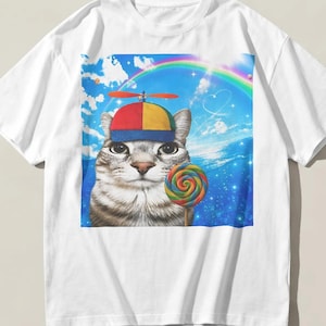 Pode incluir: Uma t-shirt branca de manga curta com uma estampa gráfica de um gato malhado usando um boné de hélice multicolorido. O gato segura um pirulito colorido em espiral contra um fundo de céu azul com um arco-íris e nuvens brancas dispersas.