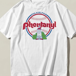 Könnte beinhalten: Ein weißes T-Shirt mit kurzen Ärmeln und einem Grafikdruck auf der Brust. Das Design zeigt ein rot-blaues Baseball-Motiv mit dem Schriftzug "PHILADELPHIA" und "Phentanyl" in roten Buchstaben über einer Abbildung eines Gebäudes und Bäumen.