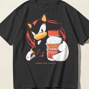 以下が含まれることがあります： 黒の半袖Tシャツ。タイレノール・エクストラ・ストレングスのボトルを持ったシャドウ・ザ・ヘッジホッグのキャラクターが描かれています。キャラクターの下には、円形の背景を背に赤い大文字で「COME AND TAKE IT」というテキストがプリントされています。