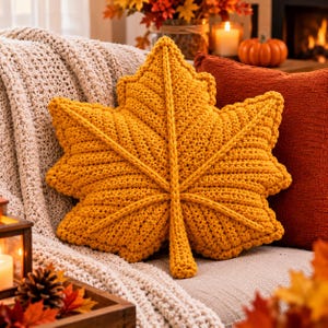 May include: A mustard yellow crochet pillow shaped like a large maple leaf. The pillow features a textured, hand-knitted surface with prominent raised veins radiating from the center stem to the pointed edges of the leaf-shaped cushion.