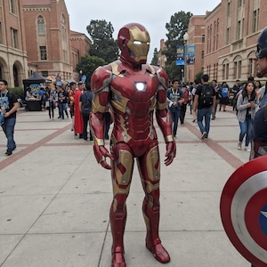 May include: A full-body Iron Man costume featuring a metallic red and gold armored suit with a helmet, glowing white eye lenses, and a circular glowing white arc reactor on the chest, standing on a paved walkway in an outdoor setting.