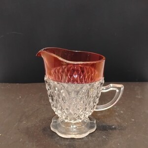 May include: A clear glass creamer pitcher featuring a diamond-patterned texture on the body and a scalloped base. The upper rim of the pitcher is finished with a deep, metallic red iridescent band, and it includes a single clear glass handle.