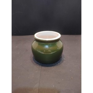 May include: A small, round ceramic pot with a glossy olive green exterior and a smooth white interior rim. The vessel features a wide, open top and a curved, bulbous body, presenting a simple, functional design with a clean, polished finish.