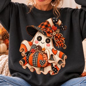 May include: A dark gray sweatshirt featuring a graphic of a ghost wearing a witch hat and holding a pumpkin. The ghost has button eyes, stitched details, and a patchwork design in orange, black, and white with plaid and polka dot patterns.