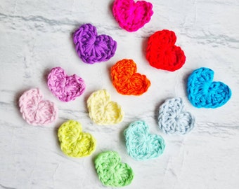 Puede incluir: Una colección de 12 corazones tejidos a crochet en varios colores, incluyendo rosa, morado, rojo, naranja, amarillo, verde, azul y azul claro. Los corazones están dispuestos en un semicírculo sobre una superficie de mármol blanco.