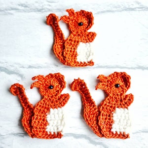 May include: Three crocheted orange squirrels with white bellies. The squirrels are sitting on a white surface.