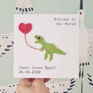 May include: A white card with a green crocheted dinosaur holding a red heart balloon. The card says "Welcome to the World" and "James Jaxon Egan! 28.06.2022".