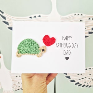 May include: A white card with a crocheted green turtle holding a red heart. The card says "Happy Father's Day Dad" with a small black heart below the text.