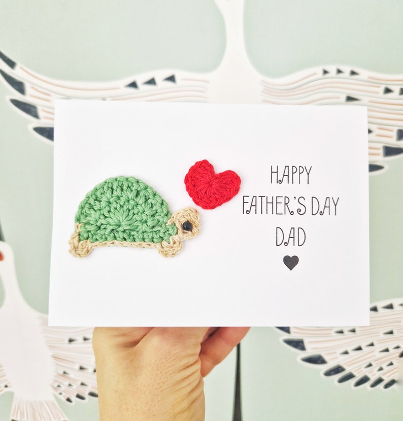 May include: A white card with a crocheted green turtle holding a red heart. The card says "Happy Father's Day Dad" with a small black heart below the text.