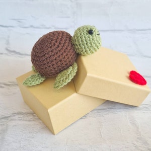 May include: A crocheted green and brown turtle toy sitting on top of two stacked brown gift boxes. The top box has a red crocheted heart on it.