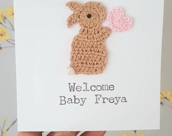 May include: A white card with a crocheted brown bunny holding a pink heart. The card says "Welcome Baby Freya".