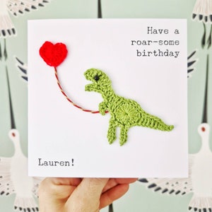 May include: A green crocheted dinosaur holding a red heart balloon with the text "Have a roar-some birthday" and "Lauren!" on a white card.