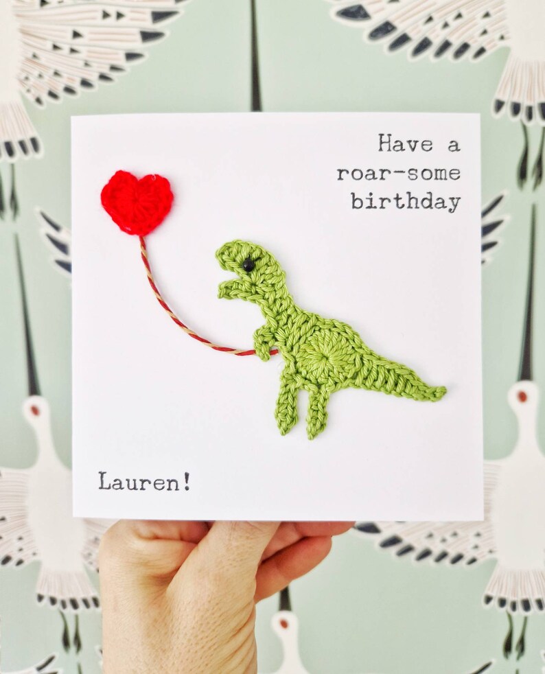 May include: A green crocheted dinosaur holding a red heart balloon with the text "Have a roar-some birthday" and "Lauren!" on a white card.