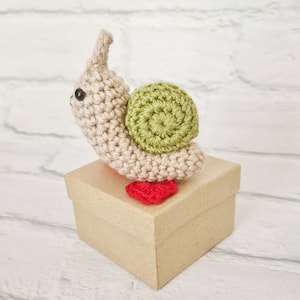 May include: A crocheted snail toy with a beige body, green shell, and red feet. The snail is sitting on a brown cardboard box.