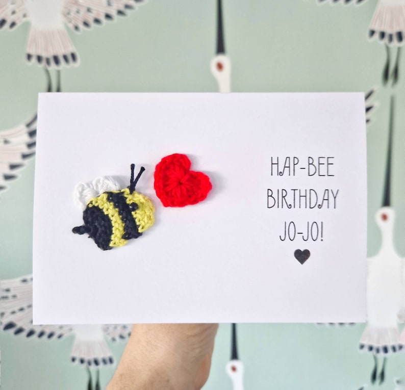 May include: A white card with a crocheted bee and red heart. The card says "Hap-Bee Birthday Jo-Jo!" with a small black heart below the text.