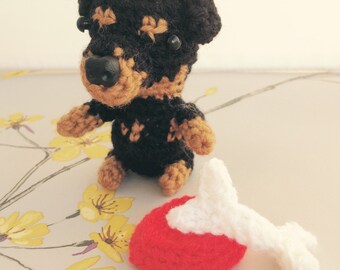Op de afbeelding: Een gehaakt Rottweiler-speelgoed met zwart en bruin haar, dat een rood en wit gehaakt bot vasthoudt.