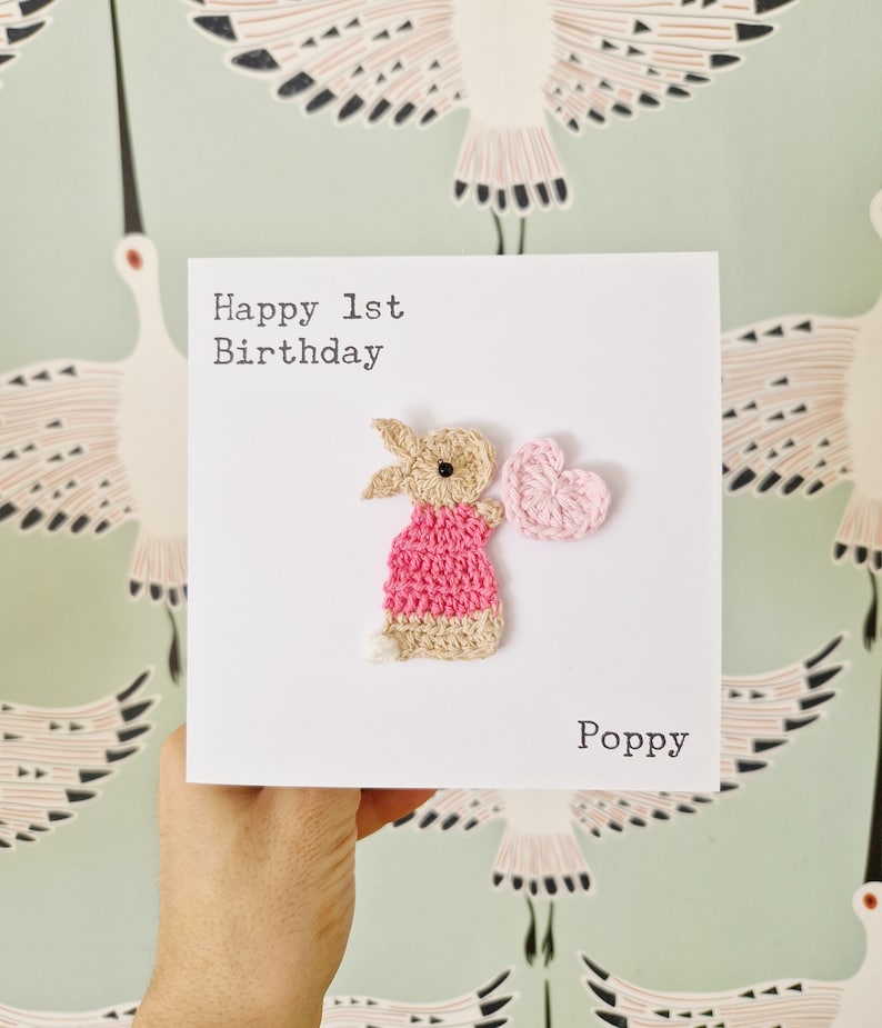 Peut inclure: Une carte faite &agrave; la main avec un lapin crochet&eacute; tenant un c&oelig;ur rose. La carte dit "Joyeux 1er anniversaire" et "Poppy".