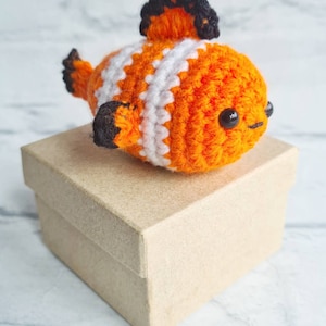 May include: A crocheted orange and white clownfish toy with black eyes and fins. The toy is sitting on a brown cardboard box.