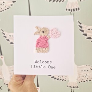 Peut inclure: Une carte faite &agrave; la main avec un lapin rose crochet&eacute; tenant un c&oelig;ur rose. La carte dit "Bienvenue Petit",