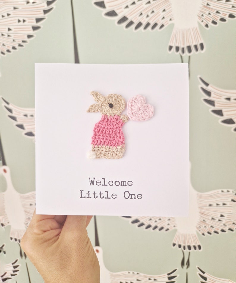 Peut inclure: Une carte faite &agrave; la main avec un lapin rose crochet&eacute; tenant un c&oelig;ur rose. La carte dit "Bienvenue Petit",