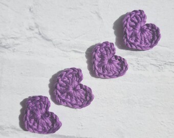 Puede incluir: Cuatro corazones de ganchillo de color morado dispuestos en fila sobre un fondo blanco.