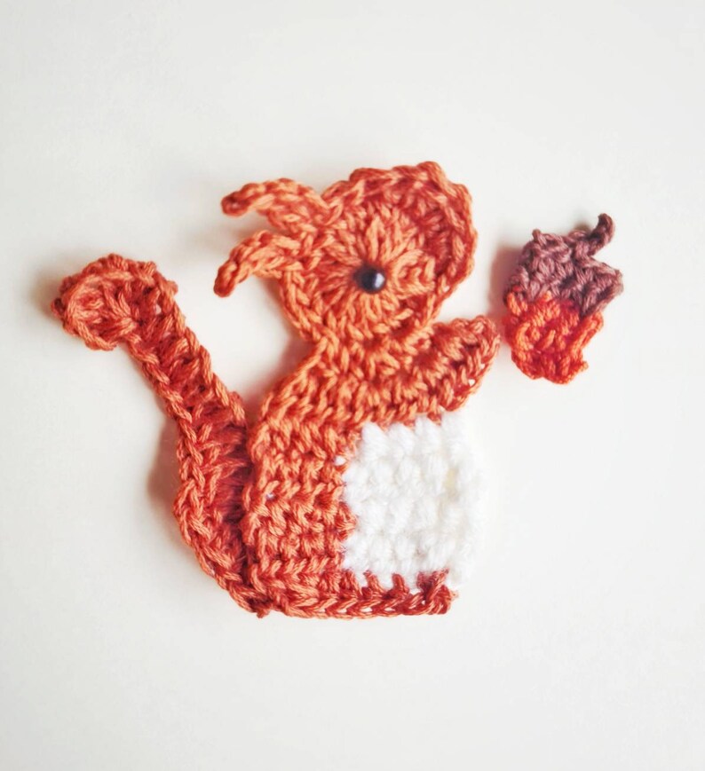 May include: A crocheted orange and white squirrel holding a brown acorn. The squirrel is made of yarn and has a white belly.