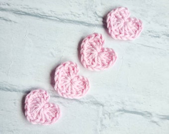 Puede incluir: Cinco corazones de ganchillo de color rosa dispuestos en fila sobre una superficie blanca.