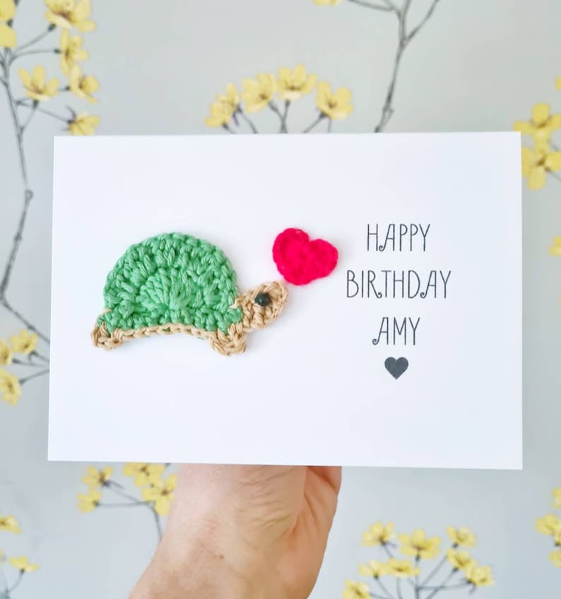 May include: A white card with a crocheted green turtle holding a pink heart. The card says "Happy Birthday Amy" with a small black heart below the name.