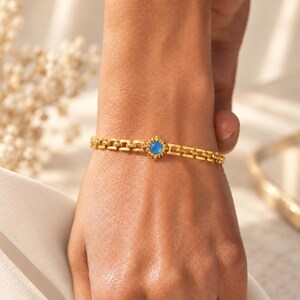 Puede incluir: Una pulsera de cadena en tono dorado con un dije central en forma de sol que presenta una piedra redonda, lisa y de color azul claro. La pulsera está formada por una serie de eslabones rectangulares texturizados que crean una banda flexible.