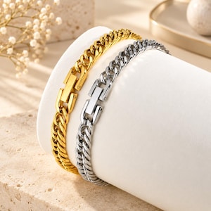 May include: Two curb chain bracelets, one in a polished gold tone and one in a polished silver tone. Each bracelet features a flat, rectangular clasp mechanism integrated into the chain links, providing a secure closure for the metallic wrist jewelry.