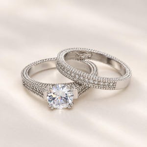 Könnte beinhalten: Ein silberfarbenes Verlobungsring-Set mit einem großen, rund geschliffenen Mittelstein, der von vier Krappen gehalten wird. Sowohl der Verlobungsring als auch der passende Ring sind mit kleinen Pavé-Steinen besetzt und haben einen strukturierten Rand.