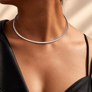Puede incluir: Un collar tipo gargantilla rígido de metal plateado, liso y pulido, con un diseño circular que se ajusta a la base del cuello. La pieza presenta una banda continua de superficie reflectante sin cierres ni adornos visibles.